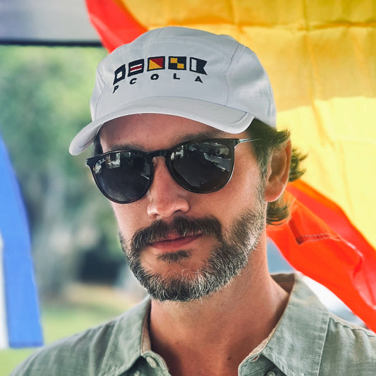 White athletic dry-fit hat featuring nautical signal flags that spell out 'PCOLA,' representing Pensacola, Florida.